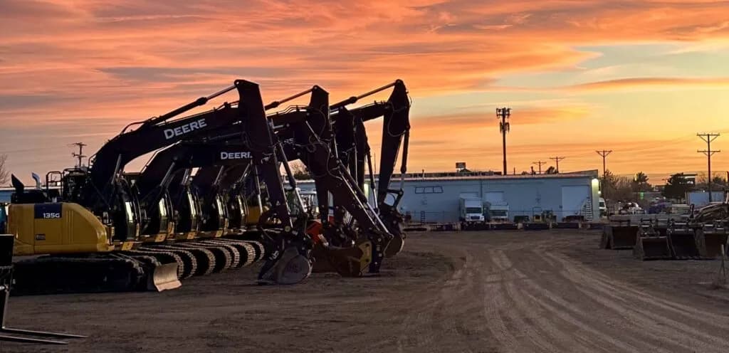 4RE | Multiple John Deere excavators in a shop yard with a sunset in the background.